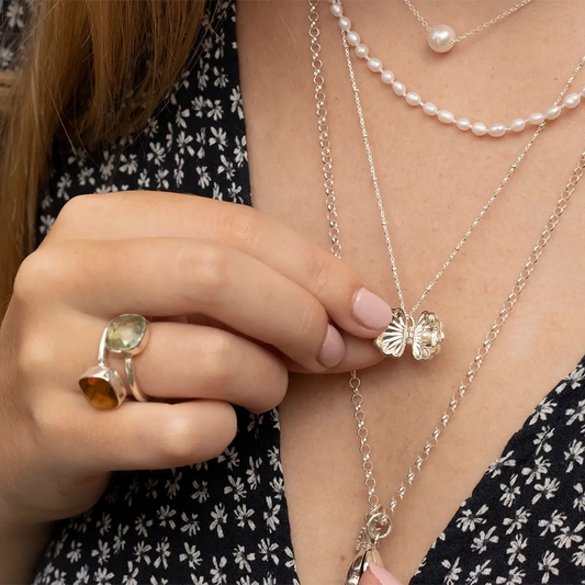 Close-up of a woman opening a sterling silver shell necklace while wearing layered pearl and silver chains and gemstone rings.