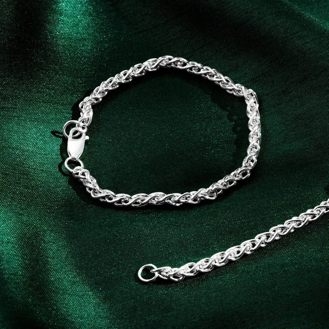 Silver spiga bracelet with a lobster clasp shown next to the end of a matching chain on green satin fabric.