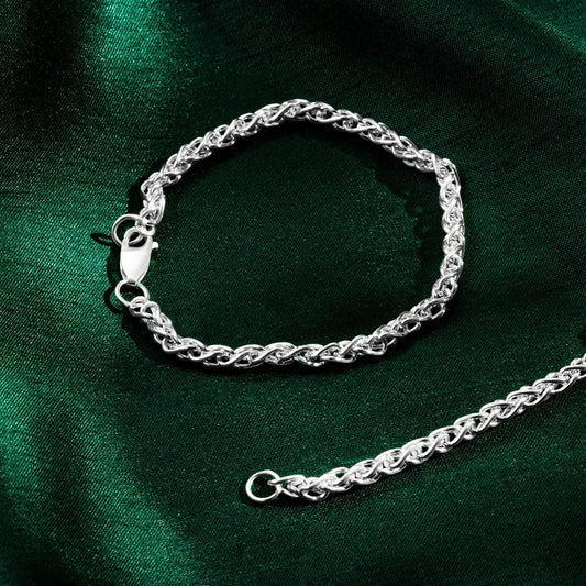 Silver spiga bracelet with a lobster clasp shown next to the end of a matching chain on green satin fabric.