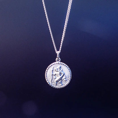 A silver round pendant necklace featuring a raised depiction of Saint Christopher carrying a child, hanging against a dark blue gradient background.