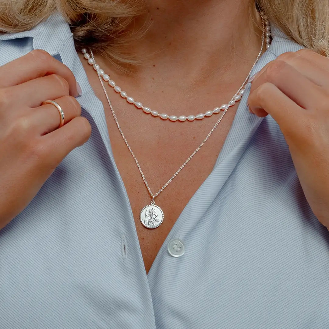 Close-up of a woman wearing a sterling silver St Christopher pendant necklace layered with a delicate freshwater pearl choker, styled with a light blue shirt.