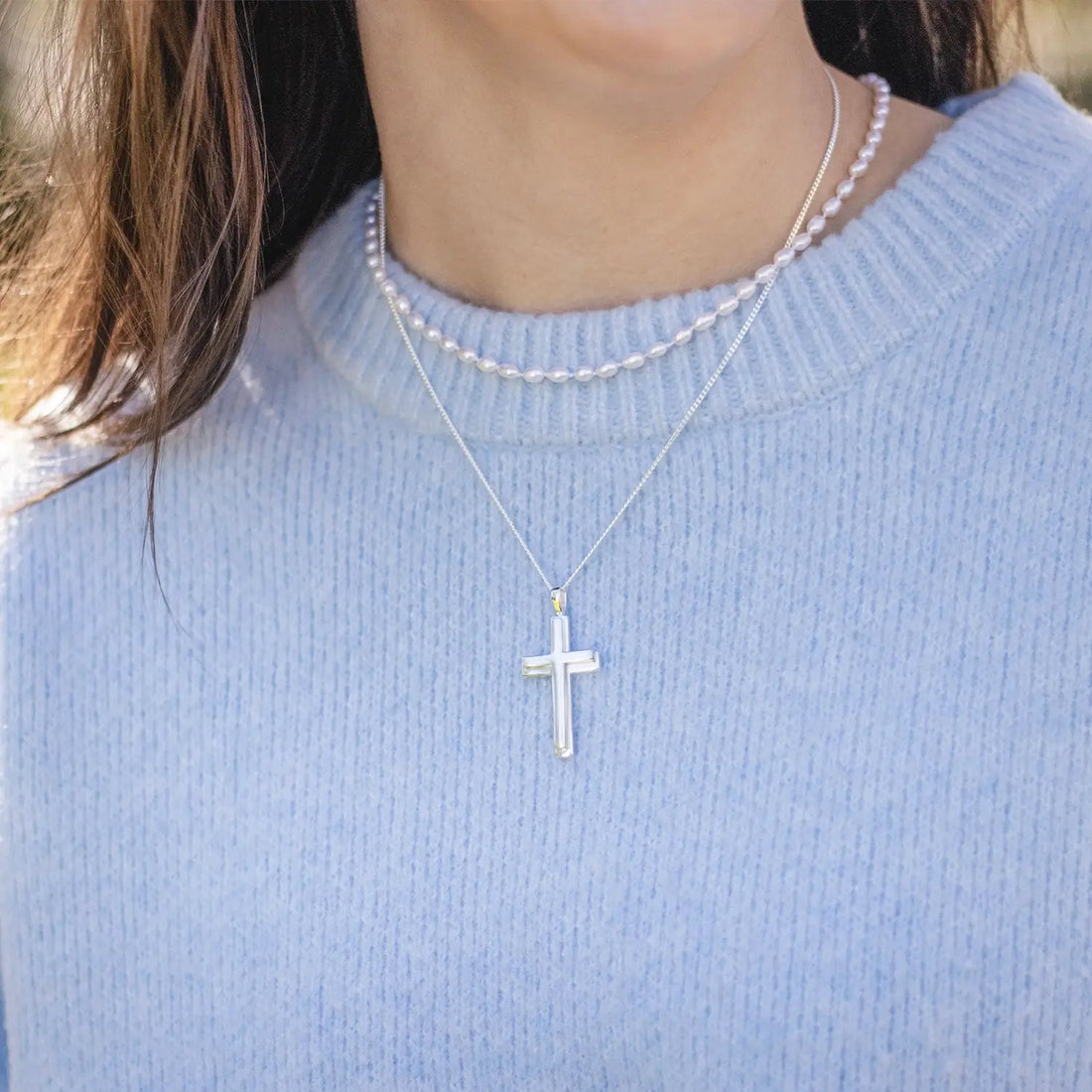 Model in a pale blue jumper wearing the Lily Blanche sterling silver Heavy Cross Necklace on a 20 inch curb chain along with a rope of ivory seed pearls 