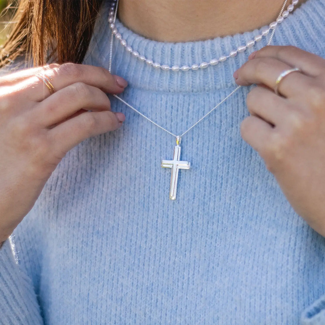 Model in a pale blue jumper wearing the Lily Blanche sterling silver Heavy Cross Necklace on a 20 inch curb chain along with a rope of ivory seed pearls 