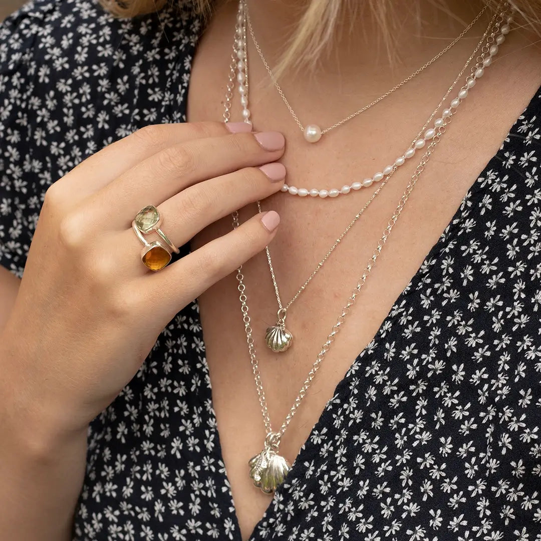 Close-up of layered sterling silver necklaces featuring a shell locket and pearl necklace, styled with gemstone rings and a floral dress.
