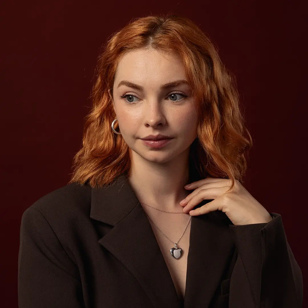 A woman with wavy red hair wearing a dark brown blazer and a solid white gold heart locket necklace with a diamond in it's centre, posing against a solid dark red background.
