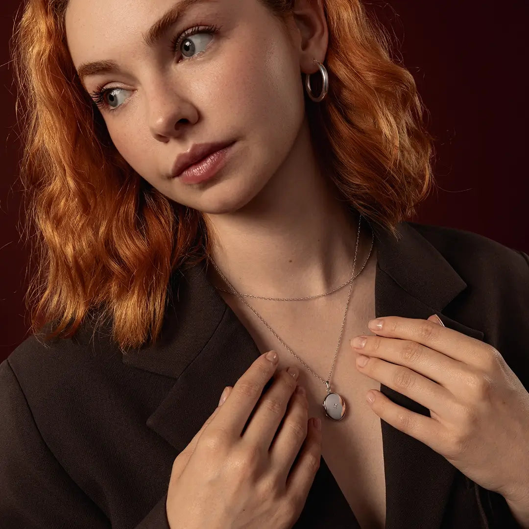 A close-up lifestyle shot of a woman with red hair wearing a delicate silver necklace with a solid white gold oval locket with a diamond in it's centre, styled with a layered white gold chain necklace and hoop earrings.