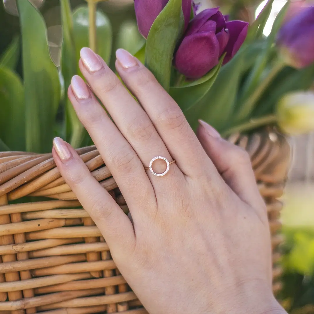 Hand wearing a 9ct solid gold open circle diamond ring, featuring a sparkling pavé-set diamond halo, resting on a wicker basket with purple tulips in the background.