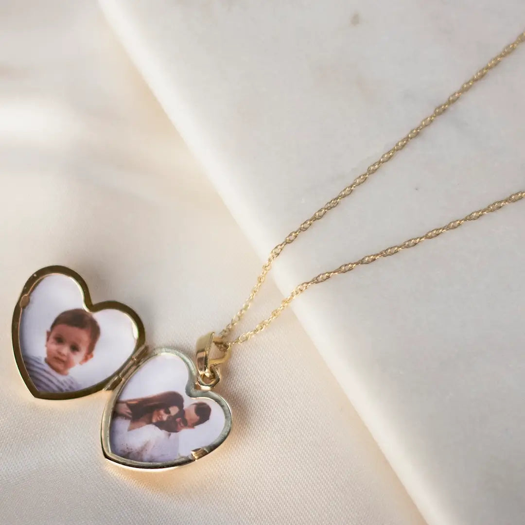 Gold heart photo locket necklace open on soft fabric showing two custom images inside, delicate fine chain detail