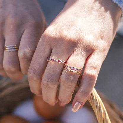 A close-up of two hands wearing gold rings; one hand features multiple gold rings with ruby and diamond details, while the other wears a simple gold band with small diamonds, with a wicker basket in the background.