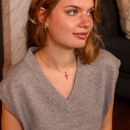Woman wearing a light grey sleeveless sweater and a gold necklace with a ruby cross pendant, sitting indoors and looking to the side