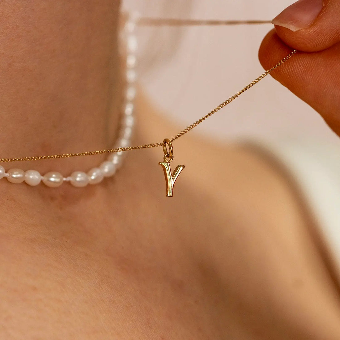 Close-up of a female model wearing a the 9-carat solid gold initial necklace, letter 'Y', on a close curb chain and a separate necklace of seed pearls. Model is holding the chain of the necklace to show it off. 