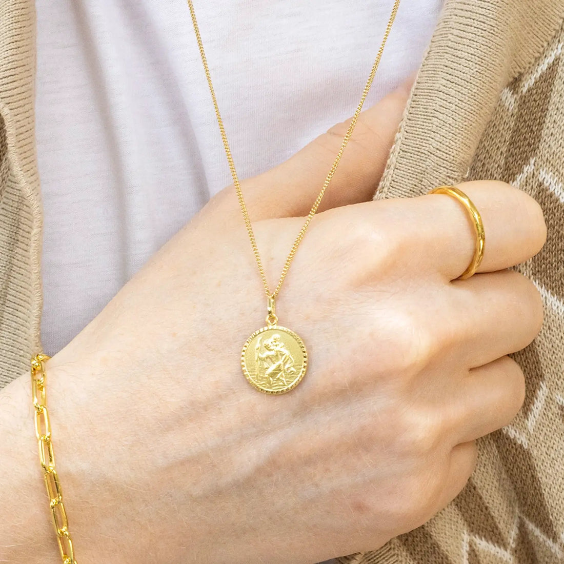 male hand wearing a gold st Christopher necklace with a round pendant, ring, and bracelet against a neutral background.