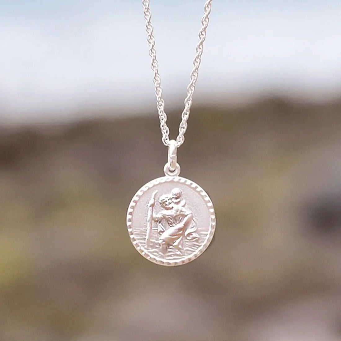 photograph of a round st Christopher necklace in silver on a curb chain, taken beside the beach and sea