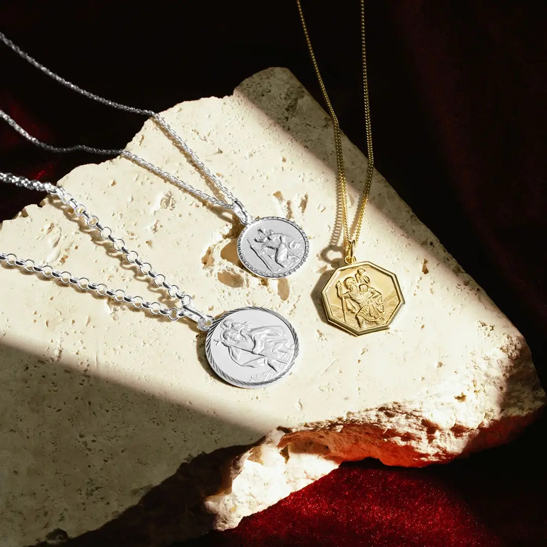 Three St. Christopher medallion necklaces displayed on a textured cream-colored stone surface. Two necklaces are silver with round medallions, each featuring a raised depiction of St. Christopher carrying a child. The third necklace is gold with an octagonal medallion featuring the same scene. The pieces are arranged with dramatic lighting and deep burgundy fabric in the background.