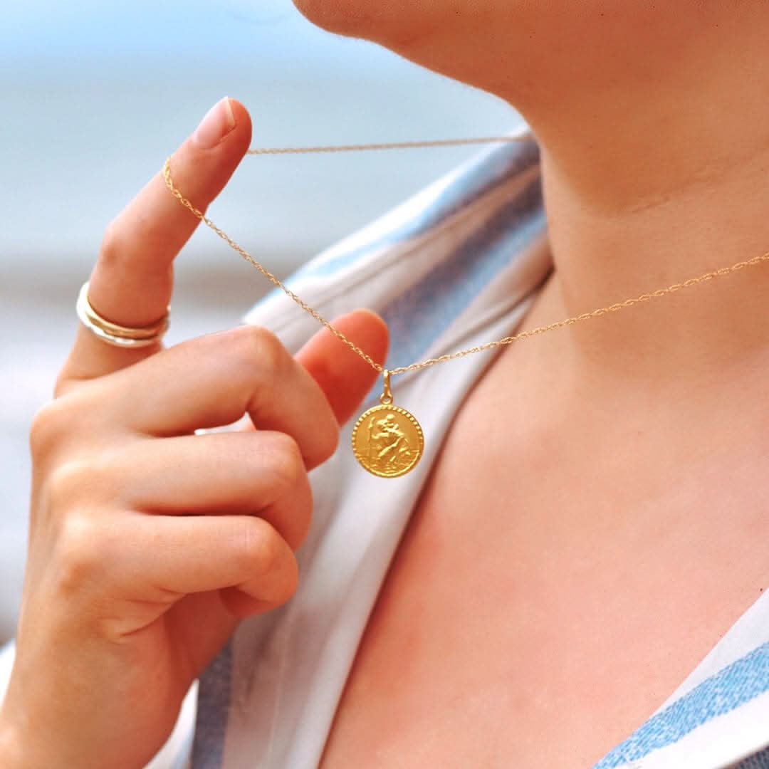Close-up lifestyle photo of a woman gently holding a gold St. Christopher necklace near her neckline. The engraved medallion pendant hangs from a fine chain, with a light, coastal background and soft lighting that evokes calm and travel inspiration