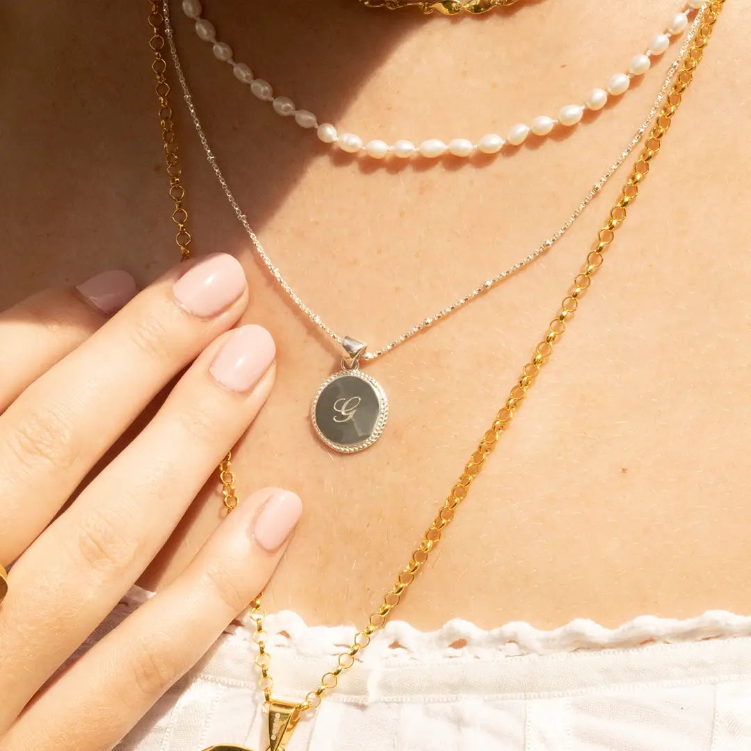Close-up of layered necklaces including a sterling silver engraved disc pendant with initial “G”, a pearl choker, and a gold chain.