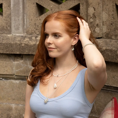 Model wearing sterling silver bird drop earrings styled with layered silver necklaces and bracelets, photographed against a stone wall background.