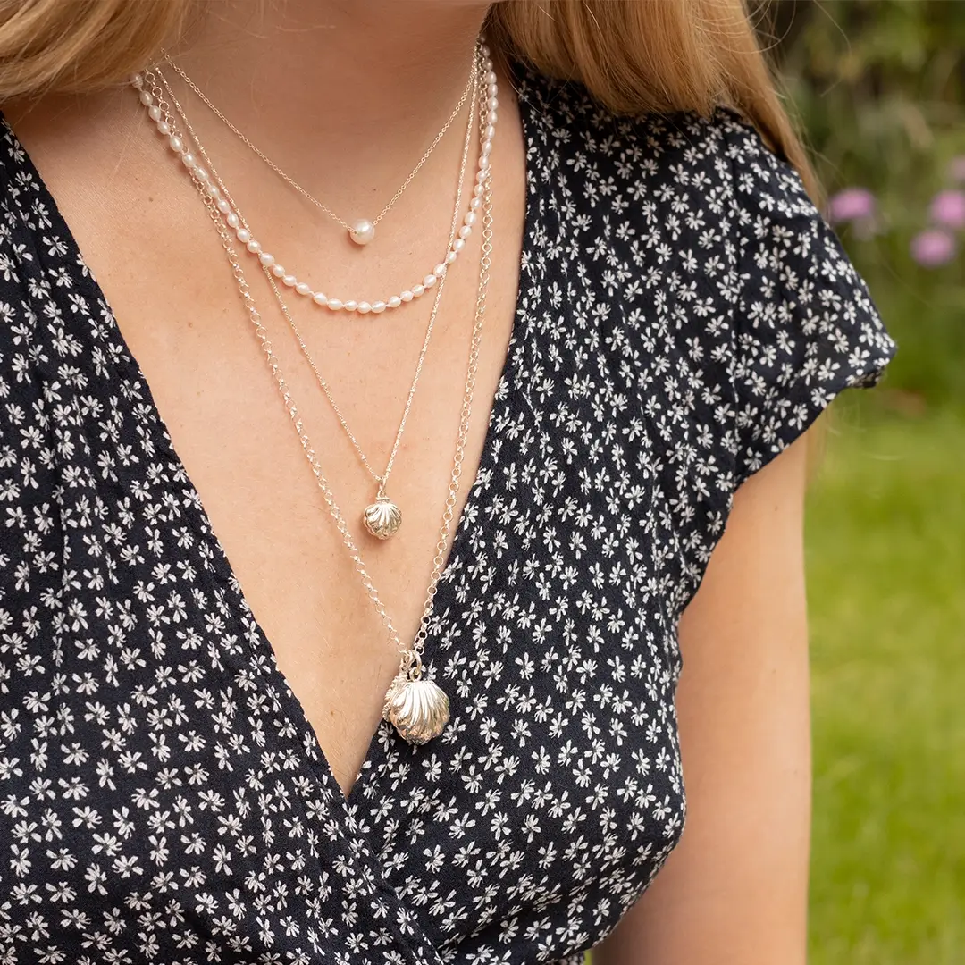 Woman wearing layered pearl and sterling silver necklaces including a scallop shell pendant and locket, styled with a black floral dress.