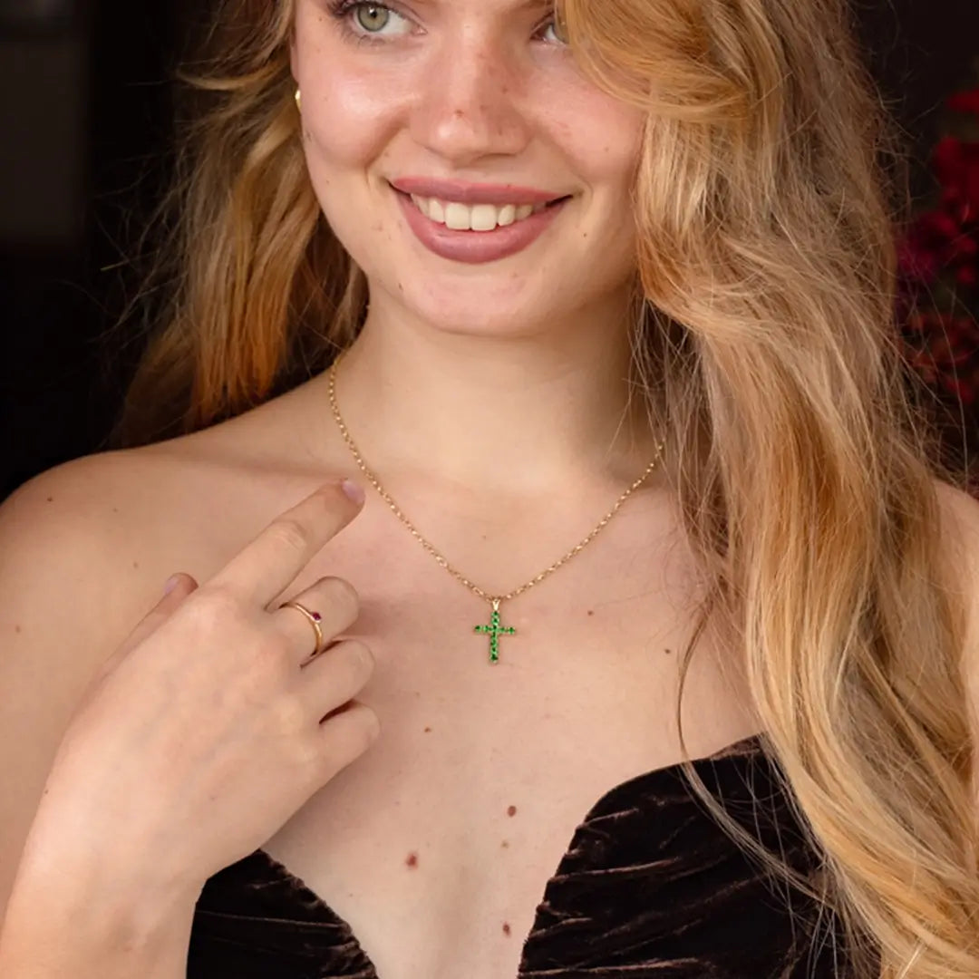 A woman with long blonde hair smiles and points to a gold necklace with a tsavorite cross pendant. She’s wearing a strapless dark velvet dress and a gold ring, with a softly lit indoor background.