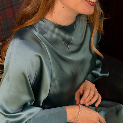 Close-up of a woman wearing a light teal satin blouse, sitting on a patterned armchair. She has a delicate white gold initial pendant necklace and a slim bracelet on her wrist.