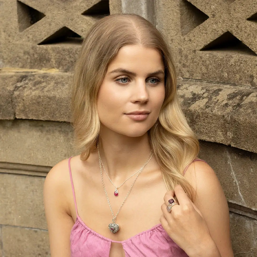 Young woman wearing a white gold heart locket necklace layered with a ruby teardrop pendant, styled with a pink dress against a stone background