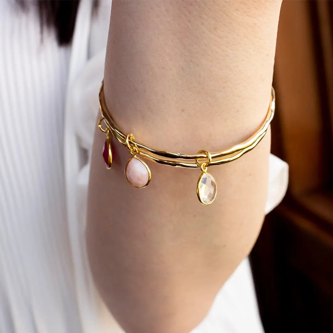 Gold charm bangle worn on wrist, featuring three faceted teardrop gemstones in soft pink, ruby red and clear white, set in polished gold with a delicate, elegant finish.