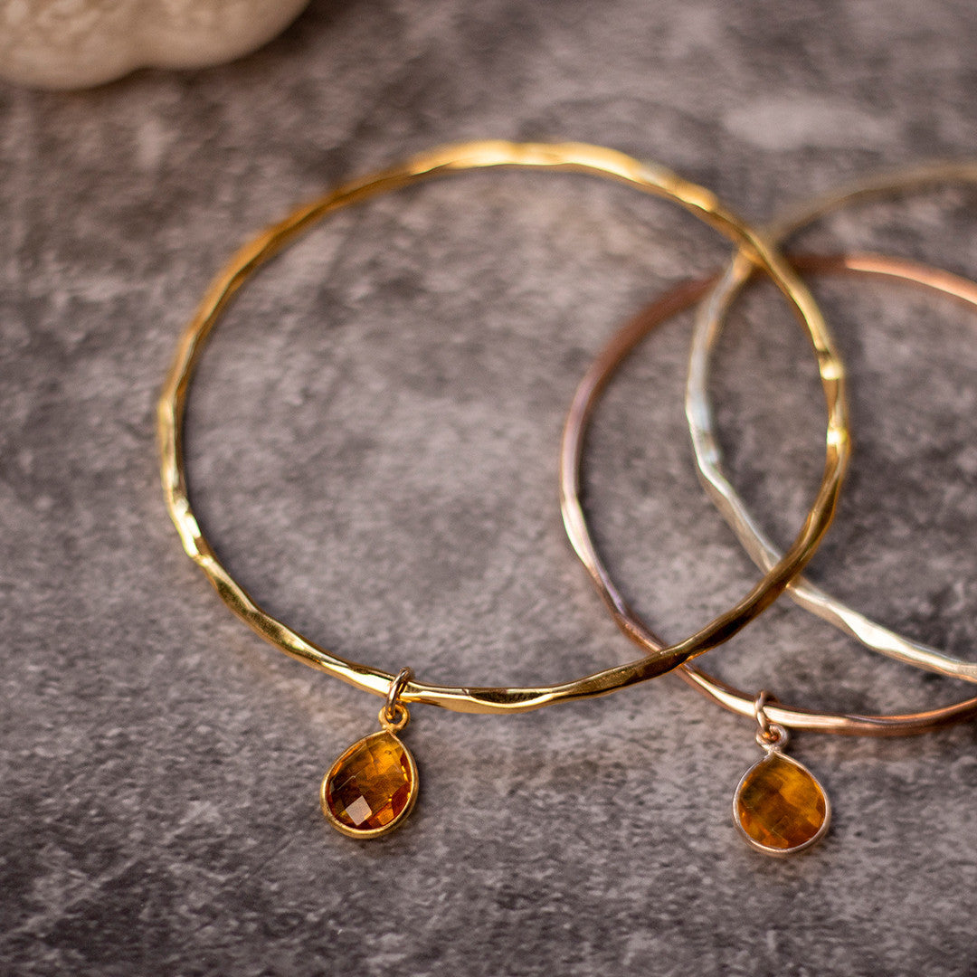 three citrine charm bangles in silver, gold and rose gold on a grey piece of fabric