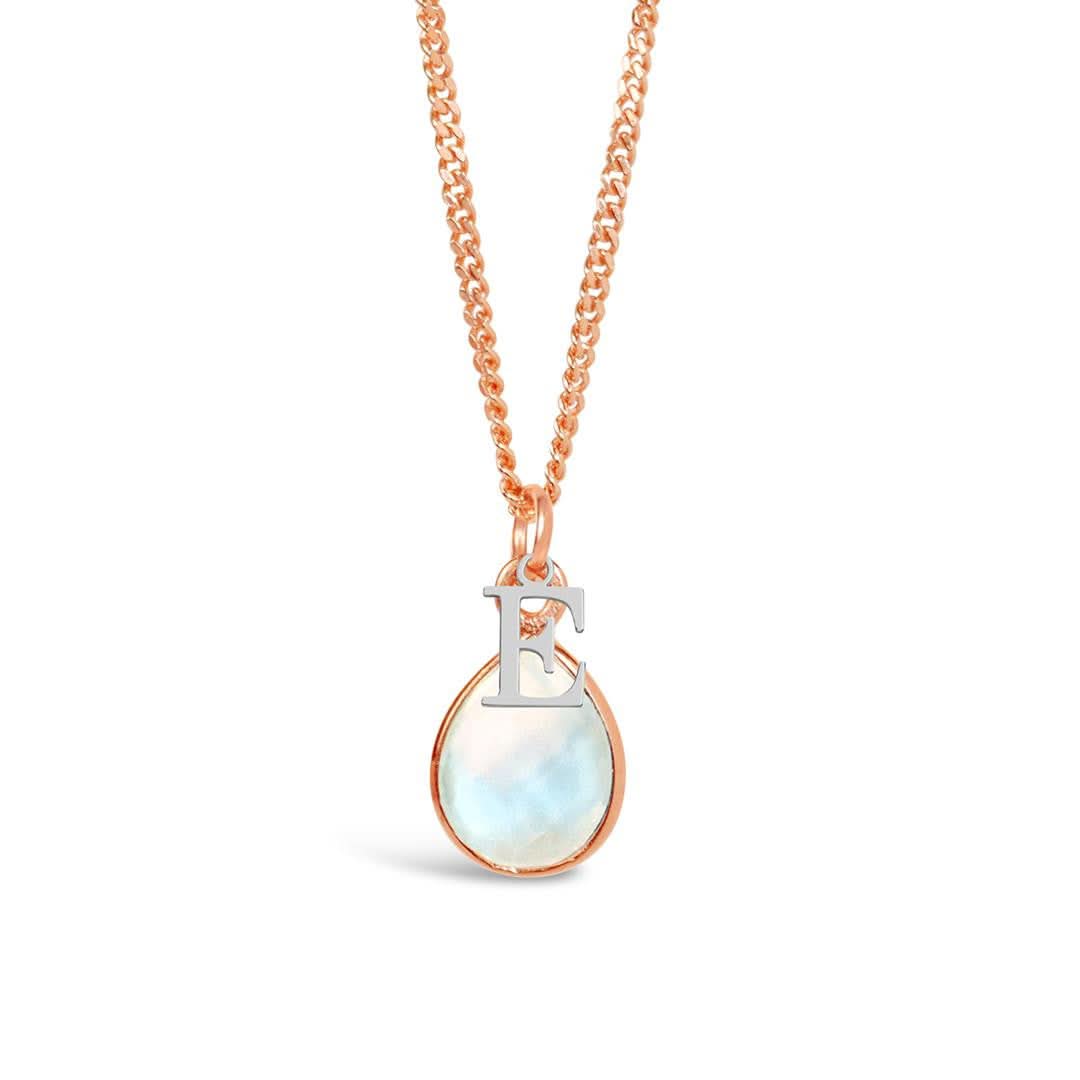 A natural moonstone charm necklace in a rose gold setting with a silver initial charm - letter E - hanging from a rose gold curb chain, photographed on a white background.