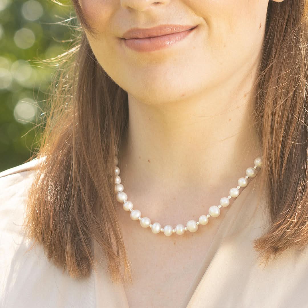 Woman wearing an ivory pearl necklace with silver spacer beads and matching pearl stud earrings, photographed outdoors in warm natural light with greenery in the background.