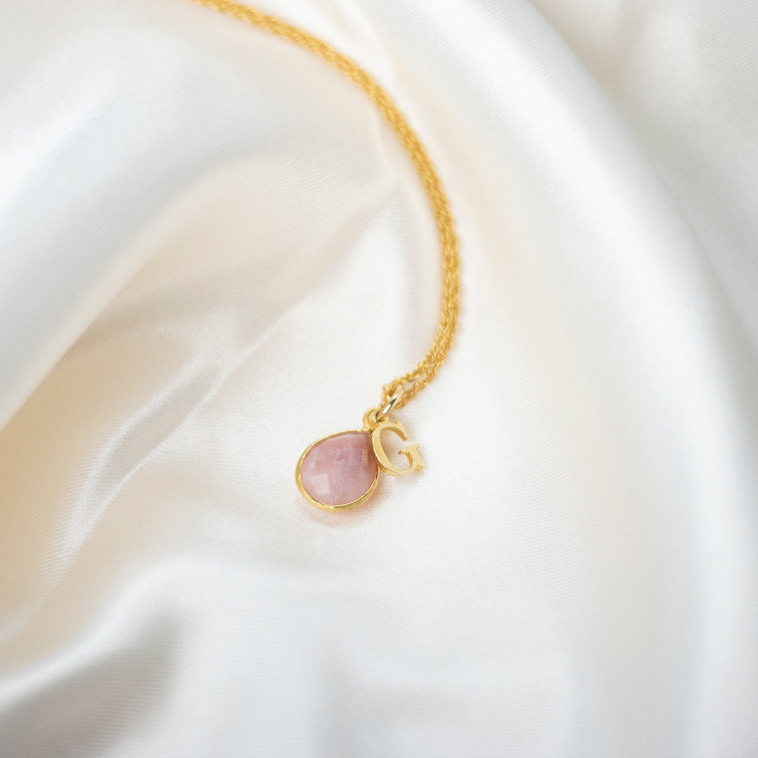 pink opal charm necklace in gold with initial charm on a piece of fabric