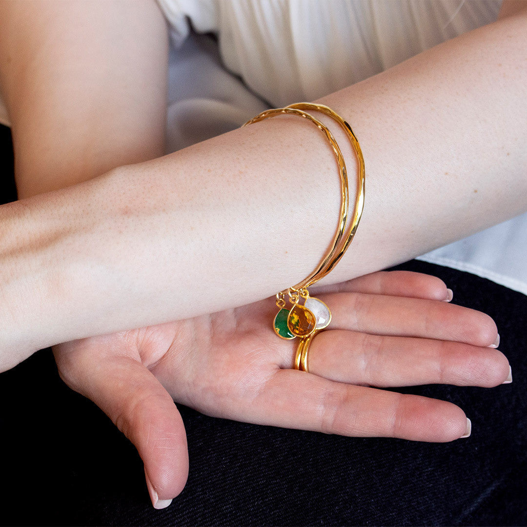 close up of model arm wearing two gold charm bangles with birthstones