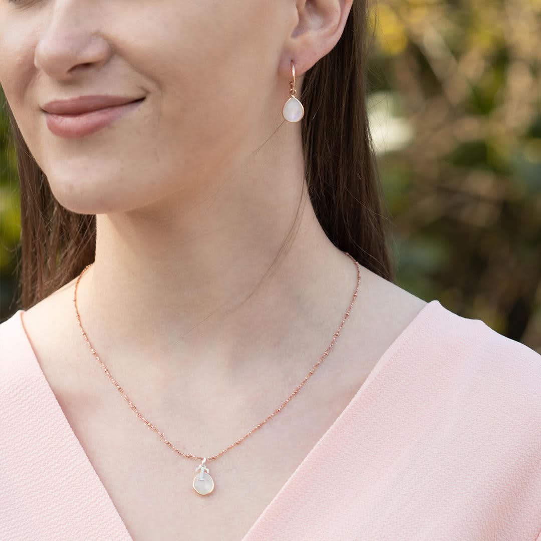 A model in a pink top, wearing moonstone drop hoop earrings in rose gold and rose gold moonstone charm necklace on a rose gold beaded chain. 