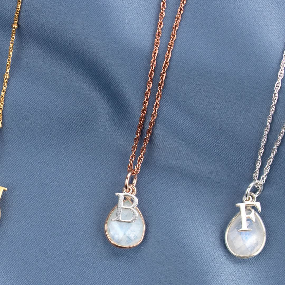 two moonstone charm necklaces in silver and rose gold  with silver initial charms photographed on blue satin fabric