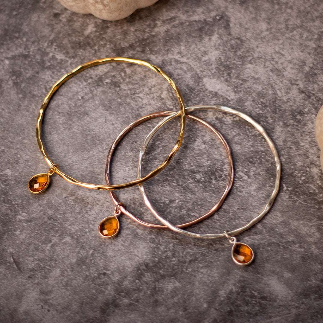 three citrine charm bangles in silver, gold and rose gold on a piece of grey fabric