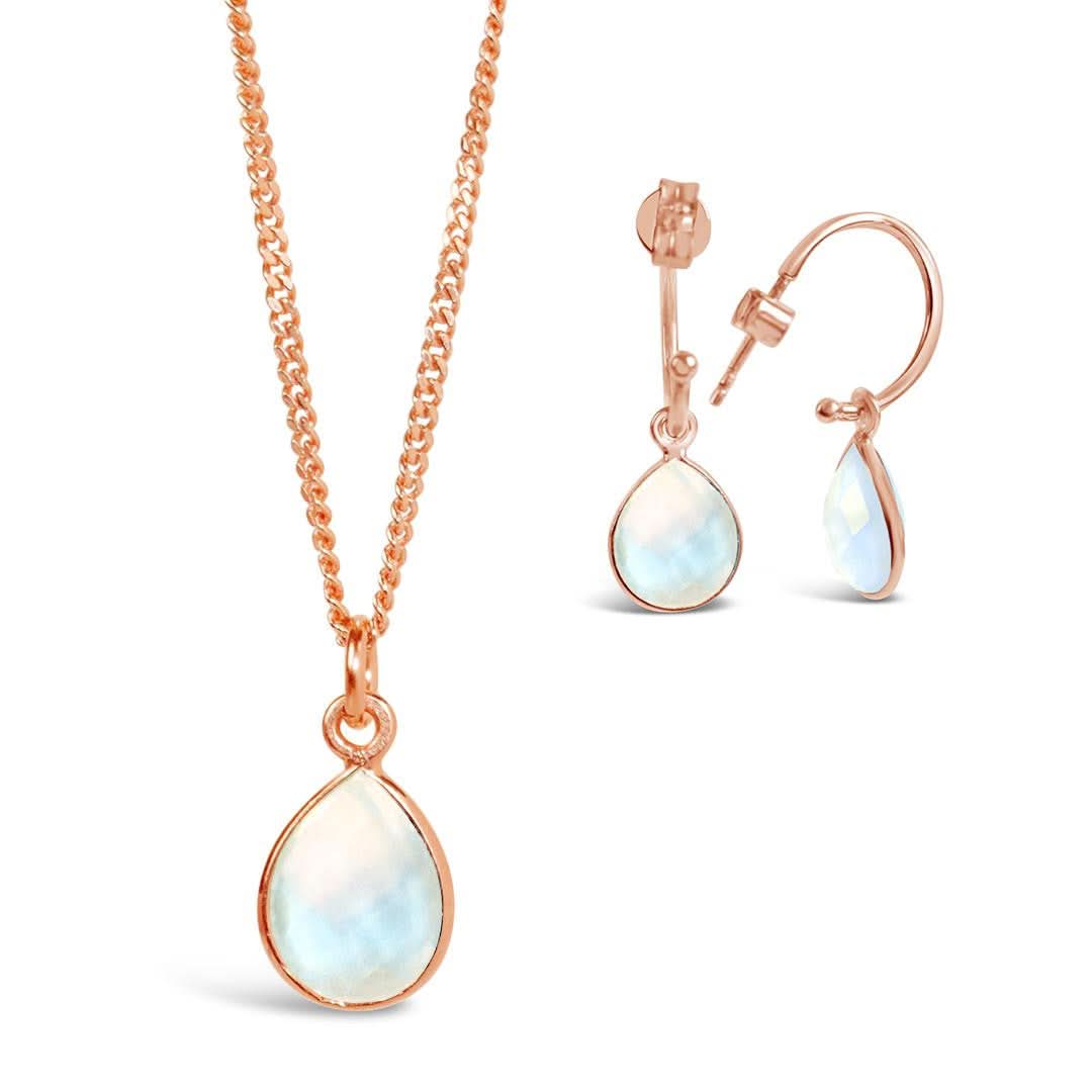 A natural moonstone charm necklace in a rose gold setting, hanging from a rose gold curb chain, along side a pair of moonstone and rose gold hoop earrings photographed on a white background.