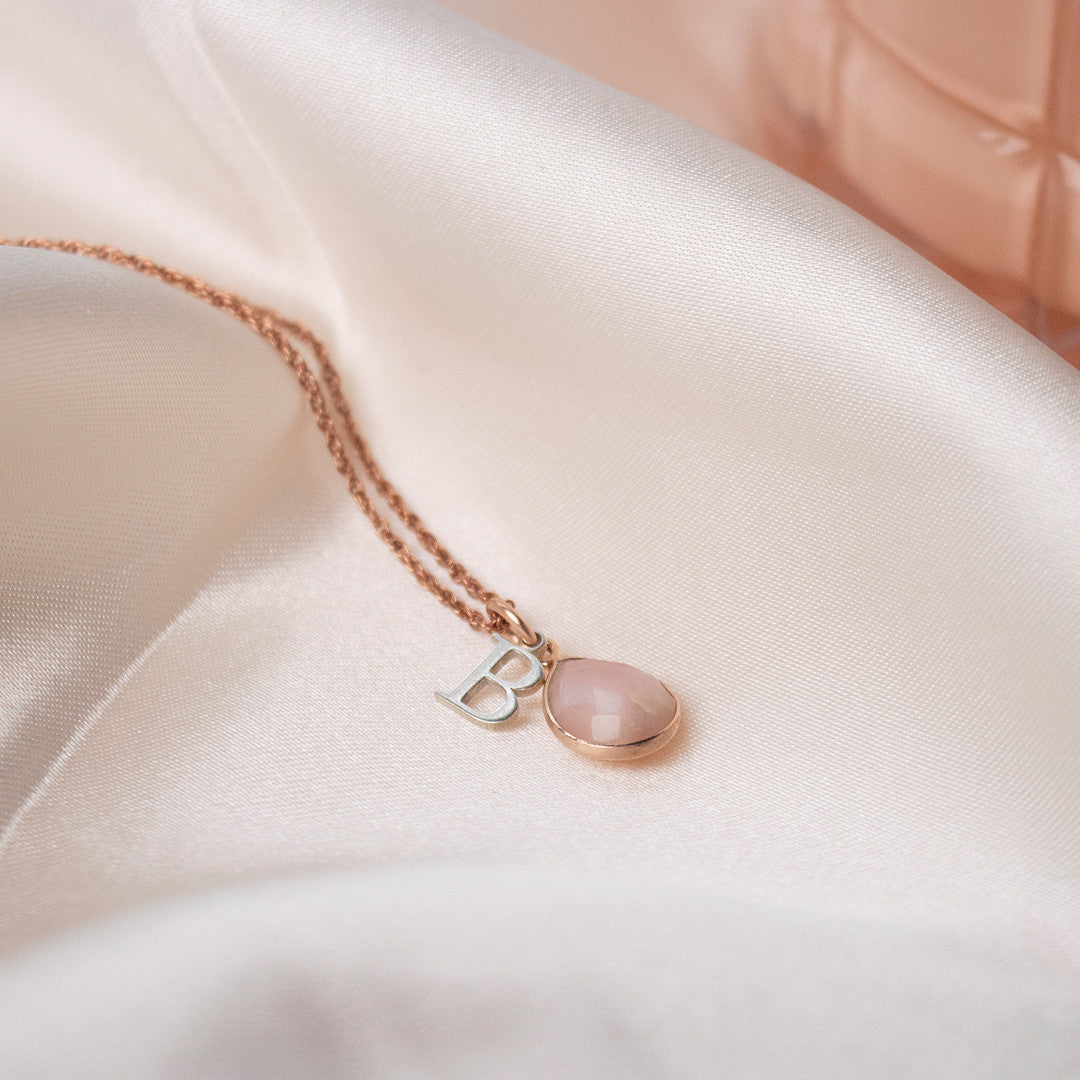 pink opal charm necklace in rose gold with silver initial charm on piece of fabric